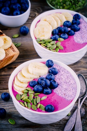 Healthy breakfast: purple smoothie bowl with chia pudding, banana, fresh blueberries and pumpkin seeds on wooden backgroundの写真素材