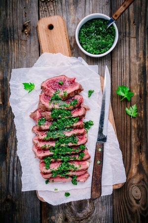 Sliced grilled barbecue beef steak with green chimichurri sauce on wooden backgroundの写真素材