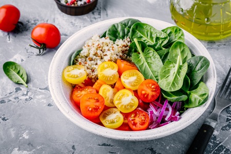Healthy green bowl salad with spinach, quinoa, yellow and red tomatoes, onions and seeds on gray backgroundの写真素材