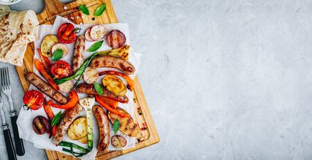 Grilled vegetables and sausages on wooden board, top view