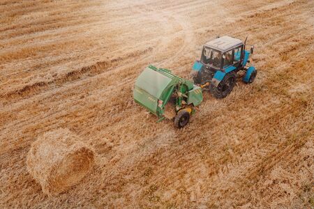 Aerial view of tractor with round baler rolling bales of straw on harvested field. View from aboveの写真素材