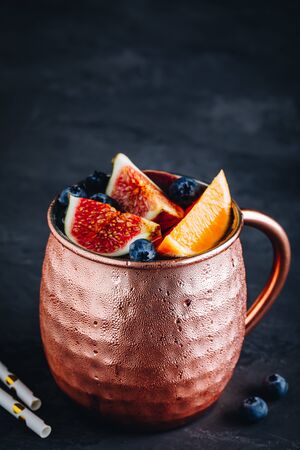 Fig moscow mule ice cold cocktail in copper cup with blueberries and orange on dark stone backgroundの写真素材