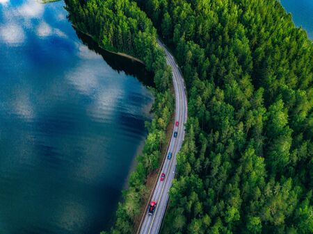 Aerial view of road with cars between green forest and blue lake water in summer Finland