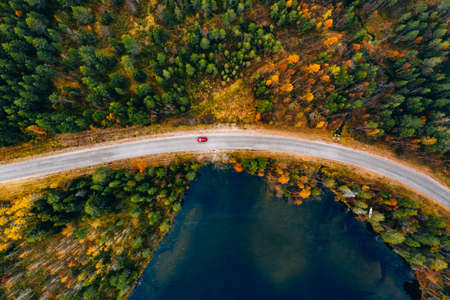Aerial view of rural road with red car in yellow and orange autumn forest with blue lake in Finland.の写真素材