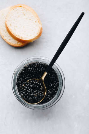 Black caviar in glass jar with spoon on white stone background, luxurious delicacy appetizer.の写真素材