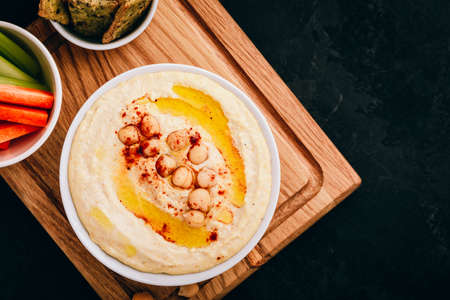 Hummus with olive oil and chickpea in bowl with vegetable sticks and crisp bread on dark stone background. Top view, with copy space.の写真素材