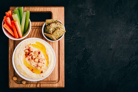 Hummus with olive oil and chickpea in bowl with vegetable sticks and crisp bread on dark stone background. Top view, with copy space.の写真素材
