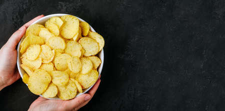 Potato chips. Woman hands holding potato chips bowl on a dark stone concrete background, top view with copy space.の写真素材