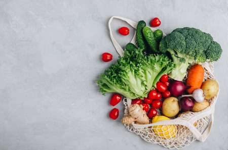 Mesh bag with vegetables. Organic broccoli, tomato, cucumbers, carrot, onions, lemon, garlic, rosemary, ginger, lettuce and potato in cotton shopper bag on gray stone background. Zero waste, plastic free concept. Healthy clean eating diet and detox. Top view with copy space.の写真素材