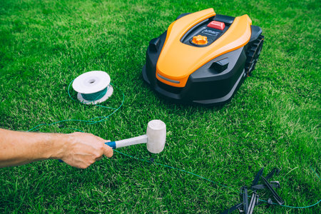 Man lays a cable and hammers in a plastic peg for autonomous operation of a robotic lawn mower. Roll of wire for robotic lawn mower, plastic pegs, connectors and robot.の写真素材