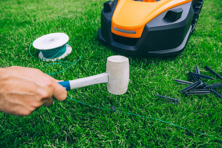 Man lays a cable and hammers in a plastic peg for autonomous operation of a robotic lawn mower. Roll of wire for robotic lawn mower, plastic pegs, connectors and robot.の写真素材