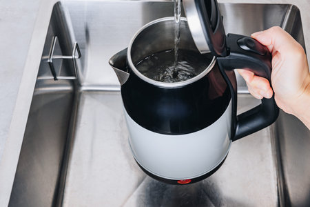 A Hand Pouring Boiling Water from an Electric Kettle into a Kitchen Sink with Metal Finish