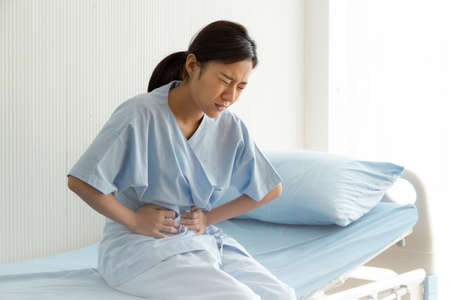 Young woman patient suffering from abdominal pain while sitting on bed at hospital. Health care...