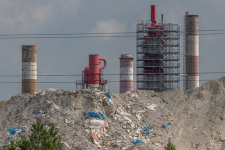 Chimneys not far from factory rise above mountains of waste. Sky is cloudy, creating heavily polluted atmosphere. Haut Rhin, Alsace, France, Europe.の写真素材