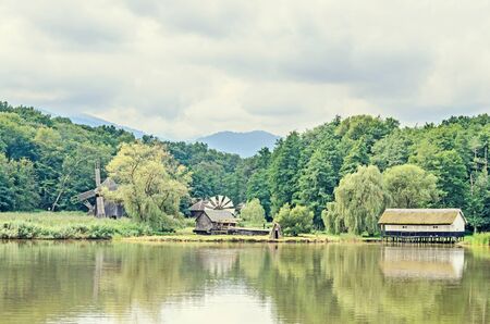 Windmill and watermill near water lake, green forest, wild vegetation, blue clouds sky.の写真素材
