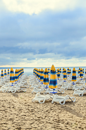 Colored stripped sun umbrellas, golden beach sand and sunbeds, sea water and clouds sky.の写真素材