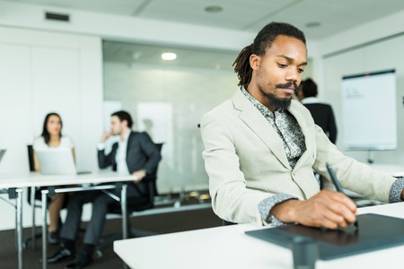 Black handsome graphics designer  with dreadlocks using digitizer in a well lit, tidy office environment  while his colleagues are working overtime in the backgroundの写真素材