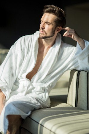 Young, muscular, handsome, healthy male in a robe relaxing on a couch in a hotel and looking towards the sunの写真素材