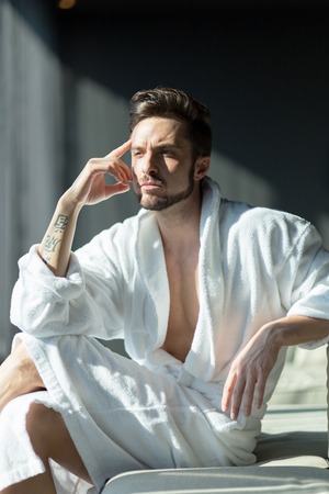 Young, handsome man in the morning thinking while sitting in a hotel room in a robeの写真素材