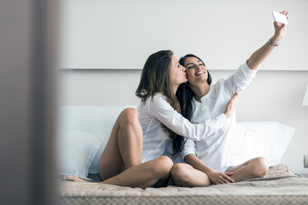Two beautiful women lying  on a bed taking a photo of themselves with a phone in a luxorious bedroom and giving a kiss on cheekの写真素材