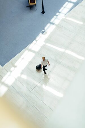 Handsome business holding a trolley and walking in a modern building seen from above