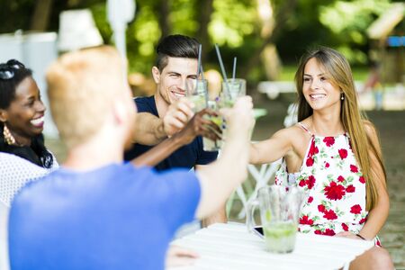 Group of cheerful happy people toasting while sitting at a table outdoors on a hot summer dayの写真素材