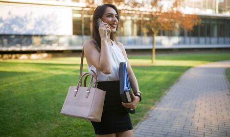 Businesswoman outdoors using phone and carrying foldersの写真素材