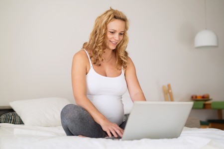 Beautiful pregnant woman using laptop on bedの写真素材