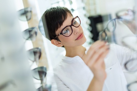 health care, eyesight and vision concept - happy beautiful woman choosing glasses at optics store