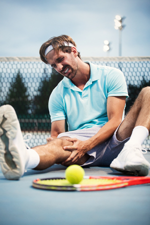 Sports injury. Young tennis player touching his knee while sitting on the tennis courtの写真素材