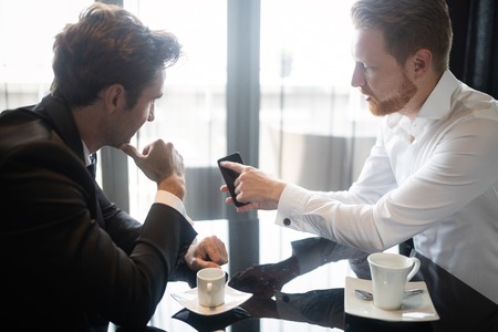 Two cheerful business people in formalwear discussing something and one of them pointing mobile