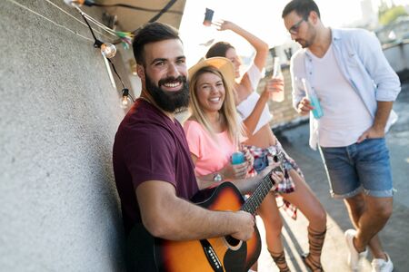 Young friends having fun at a rooftop party, playing the guitar, singing, dancing and drinking