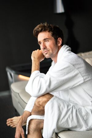 Young, handsome man in the morning thinking while sitting in a hotel room in a robeの写真素材