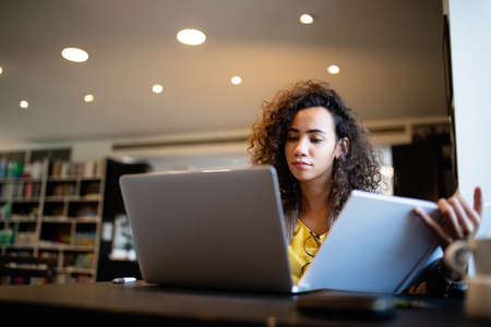 Young afro american woman sitting at table with books and laptop for finding informationの写真素材