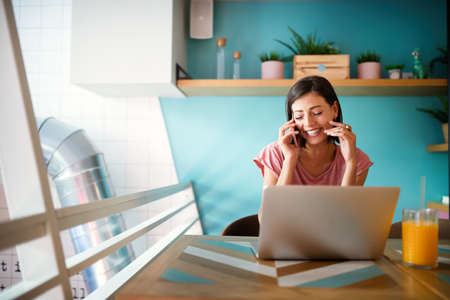 Portrait of a smiling young woman using laptop and talking on mobile phoneの写真素材