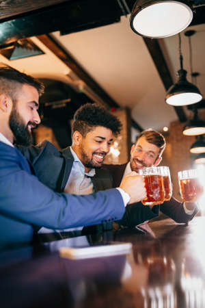 Having a pint with friend. Cheerful young men toasting with beer while sitting together at the barの写真素材