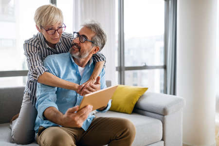 Beautiful happy mature couple is using a digital tablet, talking and smiling at homeの写真素材