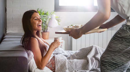 Happy man bringing the breakfast to his girlfriend on the bedの写真素材