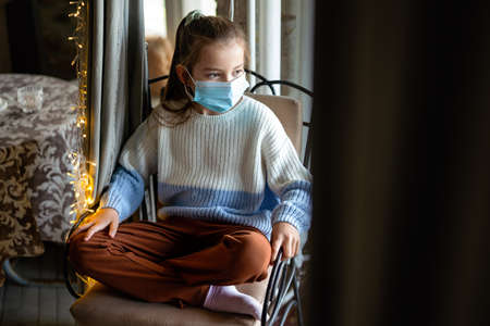Sad child girl wearing protective mask at home. People coronavirus quarantine conceptの写真素材