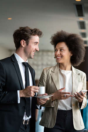 Couple of young colleagues in formal wear standing at workplace, drinking coffee near window