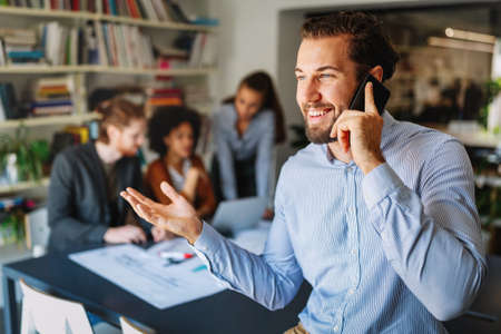 Happy young man using mobile phone and smiling while his colleagues working in officeの写真素材