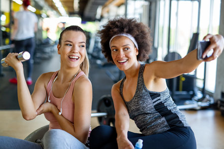Happy fit women, friends smiling, talking and taking photos after work out in gym. Social media.
