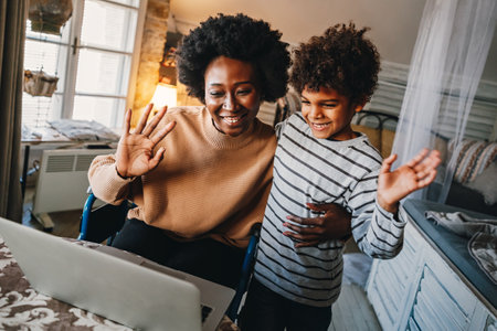 Happy african single mother with child waving hands while doing video call through laptop at homeの写真素材