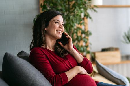 A joyful adult pregnant woman laughing and talking on her mobile phone while relaxing at homeの写真素材