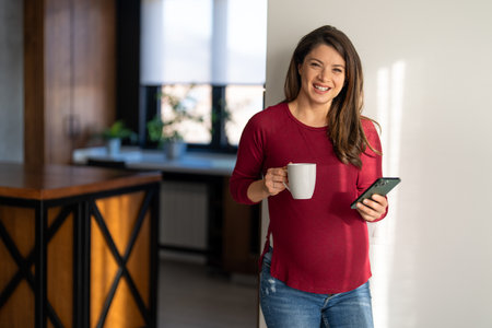 Pregnant woman enjoying and smelling cup of coffee while scrolling through her phone in the morningの写真素材