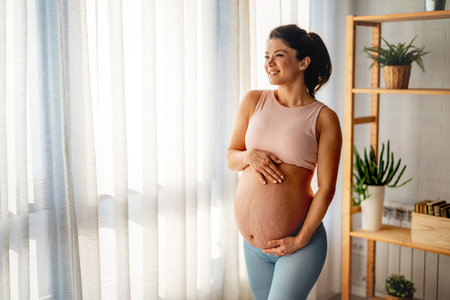 Happy fit pregnant woman in sports clothing holding stomach lovingly while standing in living roomの写真素材