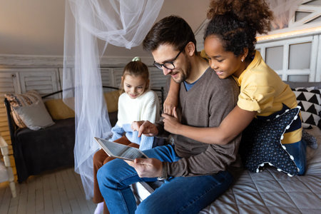 Happy diverse family resting at home on bed spending time together and using digital gadgets.の写真素材