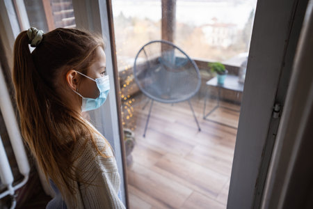 Young girl wearing face mask at home looking out of windowの写真素材
