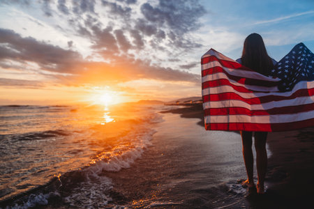 Beautiful patriotic young woman with the American flag held in her standing in the summer sunsetの写真素材