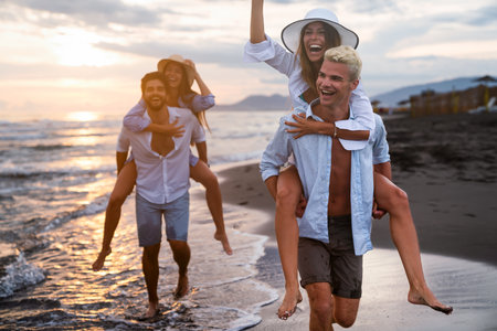 Group of happy friends, boyfriends piggybacking their girlfriends at sunset on beachの写真素材